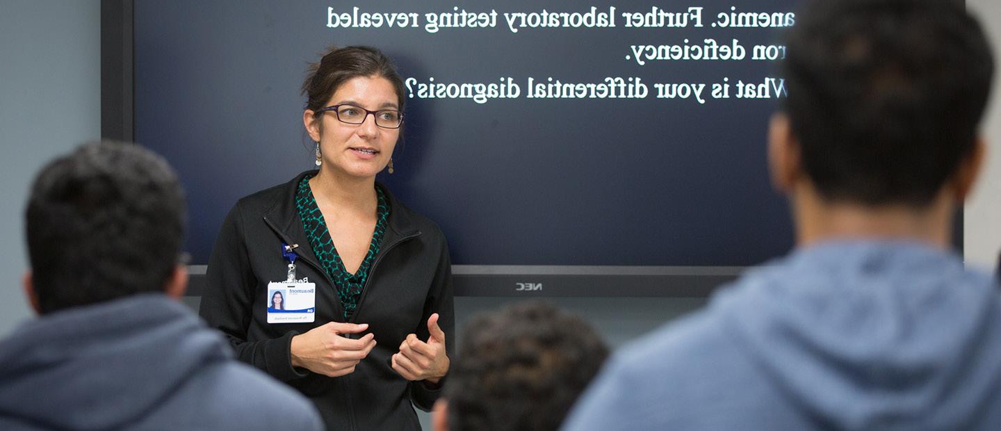 A woman wearing an ID badge, standing in front of a screen, presenting to a group.